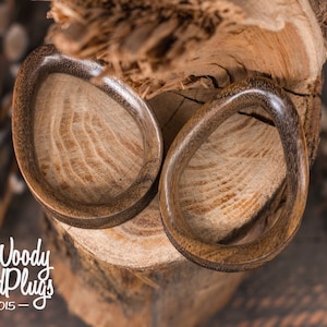 May include: Two brown wooden ear gauges, one on top of the other, with a natural wood grain pattern. The gauges are on a wooden surface.
