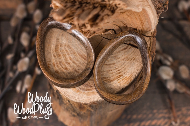 May include: Two brown wooden ear gauges, one on top of the other, with a natural wood grain pattern. The gauges are on a wooden surface.