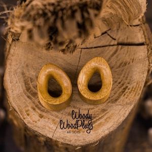 May include: Two wooden ear gauges, shaped like an oval with a teardrop-shaped hole in the center. The gauges are a light brown color and are sitting on a wooden surface. The text "Woody WoodPlugs - est. 2015" is visible below the gauges.