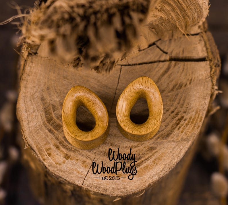 May include: Two wooden ear gauges, shaped like an oval with a teardrop-shaped hole in the center. The gauges are a light brown color and are sitting on a wooden surface. The text "Woody WoodPlugs - est. 2015" is visible below the gauges.