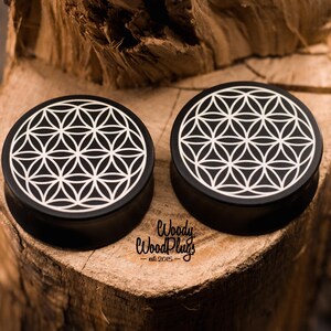 May include: Two black ear plugs with a white geometric flower of life design. The plugs are circular and sit on a wooden surface. The text "Woody Wood Plugs - est. 2015" is visible.