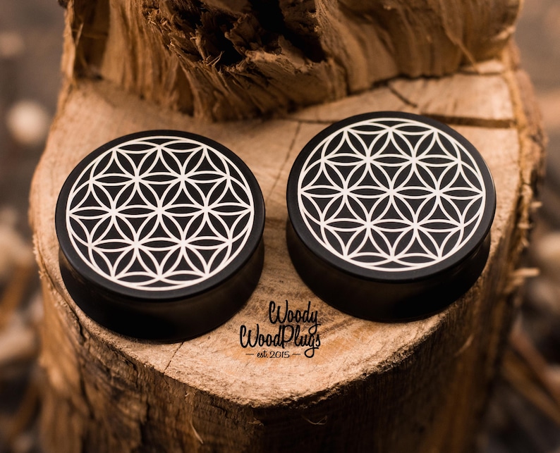 May include: Two black ear plugs with a white geometric flower of life design. The plugs are circular and sit on a wooden surface. The text "Woody Wood Plugs - est. 2015" is visible.