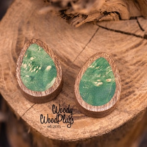 Puede incluir: Dos tapones de madera en forma de lágrima con un diseño veteado verde y blanco. Los tapones tienen un borde de madera oscura y están colocados sobre un fondo de madera natural. El texto "Woody Wood Plugs est. 2015" es visible.