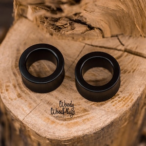 May include: Two black wooden ear gauges, one on top of the other, on a wooden surface. The ear gauges are round and have a smooth finish. The text "Woody WoodPlugs - est. 2015" is visible in the bottom center of the image.