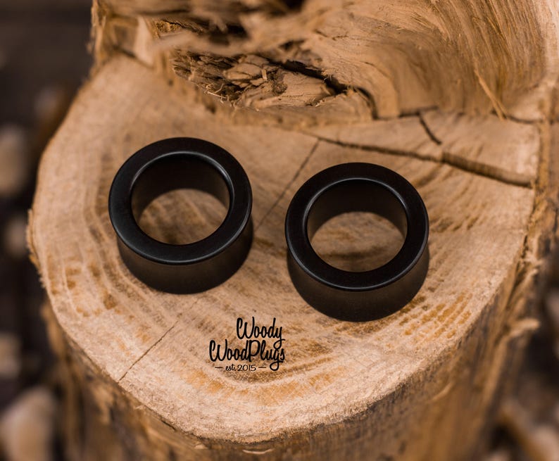 May include: Two black wooden ear gauges, one on top of the other, on a wooden surface. The ear gauges are round and have a smooth finish. The text "Woody WoodPlugs - est. 2015" is visible in the bottom center of the image.