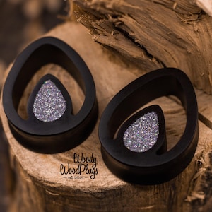 Könnte beinhalten: Zwei schwarze, tropfenförmige Ohrstöpsel mit einem glitzernden silbernen Mittelteil. Die Ohrstöpsel sind aus Holz gefertigt und haben eine glatte Oberfläche. Der Markenname "Woody WoodPlugs" ist unten rechts sichtbar.