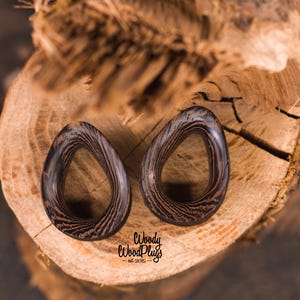 May include: Two dark brown wooden ear gauges with a striped pattern. The gauges are shaped like a teardrop with a hole in the center. The gauges are sitting on a piece of wood.