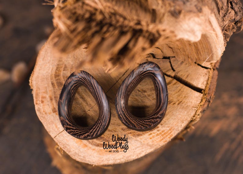 May include: Two dark brown wooden ear gauges with a striped pattern. The gauges are shaped like a teardrop with a hole in the center. The gauges are sitting on a piece of wood.