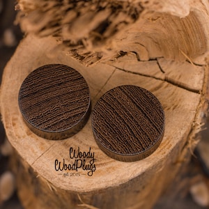 May include: Two brown wooden ear gauges with a wood grain pattern. The gauges are on a wooden surface. The text "Woody WoodPlugs - est. 2015 -" is visible below the gauges.