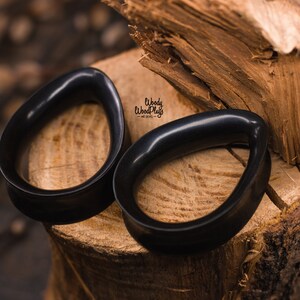 May include: Two black wooden ear gauges, shaped like teardrops, are displayed on a wooden surface. The gauges are smooth and polished, with a glossy finish.