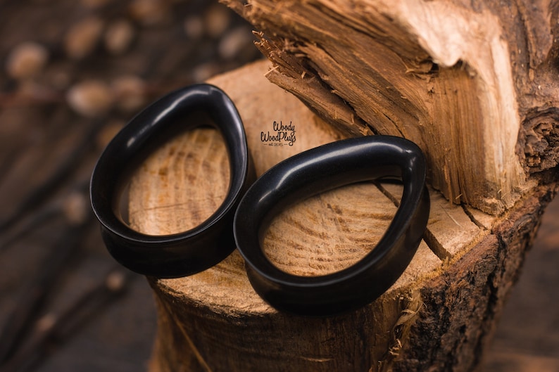 May include: Two black wooden ear gauges, shaped like teardrops, are displayed on a wooden surface. The gauges are smooth and polished, with a glossy finish.