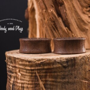 May include: Two brown wooden ear plugs on a wooden surface. The ear plugs are smooth and have a natural wood finish. The surface is a tree stump with a rough texture.