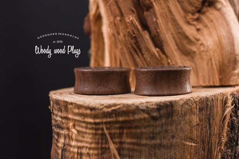May include: Two brown wooden ear plugs on a wooden surface. The ear plugs are smooth and have a natural wood finish. The surface is a tree stump with a rough texture.