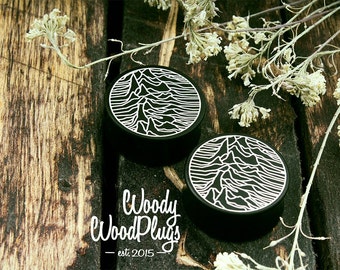 Puede incluir: Dos tapones para los oídos negros con un diseño de cordillera blanca. Los tapones son redondos y están sobre una superficie de madera rústica. El logotipo de Woody WoodPlugs es visible en la esquina inferior izquierda.
