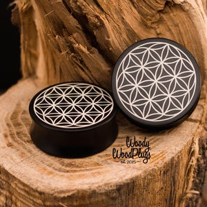May include: Two black wooden ear plugs with a white geometric flower of life design. The plugs are displayed on a piece of wood. The text "Woody Wood Plugs est. 2015" is visible.