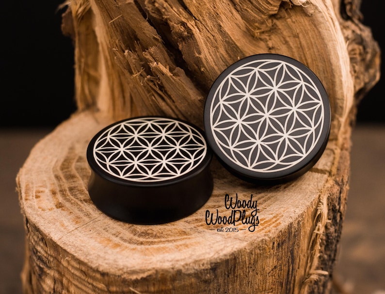 May include: Two black wooden ear plugs with a white geometric flower of life design. The plugs are displayed on a piece of wood. The text "Woody Wood Plugs est. 2015" is visible.