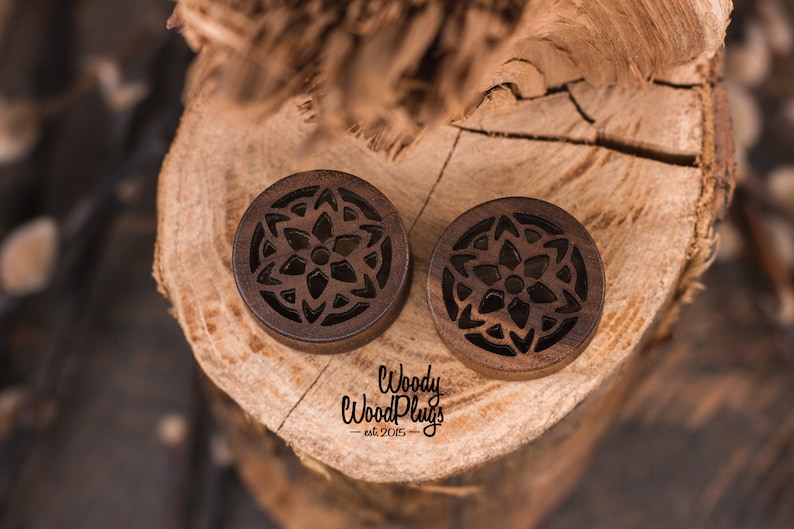 May include: Pair of brown wooden ear gauges with a carved mandala design. The gauges are on a wooden surface with the text "Woody Wood Plugs - est. 2015 -" visible.