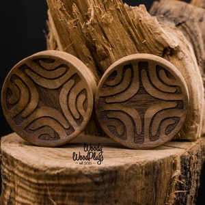 Op de afbeelding: Twee houten oorpluggen met een gegraveerd geometrisch ontwerp. De pluggen zijn bruin en hebben een natuurlijke houten afwerking. De pluggen liggen op een houten oppervlak.
