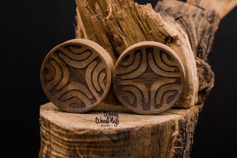 Op de afbeelding: Twee houten oorpluggen met een gegraveerd geometrisch ontwerp. De pluggen zijn bruin en hebben een natuurlijke houten afwerking. De pluggen liggen op een houten oppervlak.