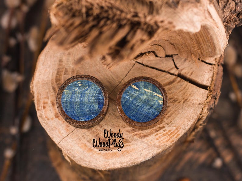 May include: Two round wooden plugs with blue and white marbled designs. The plugs are set in a dark brown wooden frame. The plugs are displayed on a wooden surface. The text "Woody WoodPlugs est. 2015" is visible.