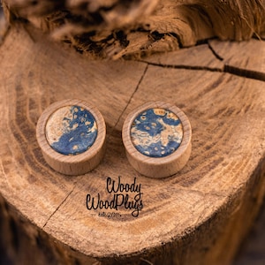 May include: A pair of wooden ear plugs with a circular blue and tan design, set against a rustic wooden backdrop. The image includes the text "Woody WoodPlugs - est. 2015".
