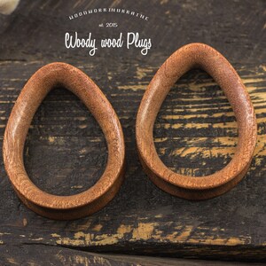 May include: Two brown wooden teardrop-shaped ear gauges on a dark wood background. The gauges are smooth and have a natural wood finish.