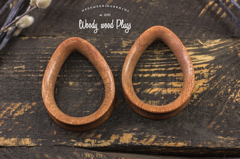 May include: Two brown wooden teardrop-shaped ear gauges on a dark wood background. The gauges are smooth and have a natural wood finish.