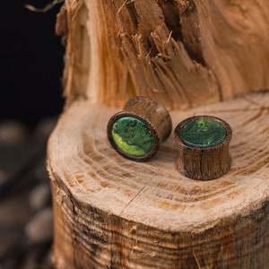 May include: Two wooden ear plugs with a green and yellow resin inlay. The plugs are cylindrical with a flared edge and sit on a textured wooden surface. The background is dark, highlighting the natural wood grain.