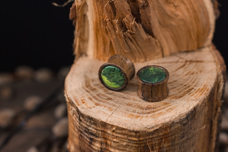 May include: Two wooden ear plugs with a green and yellow resin inlay. The plugs are cylindrical with a flared edge and sit on a textured wooden surface. The background is dark, highlighting the natural wood grain.