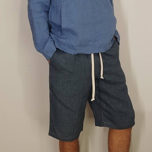 May include: A blue linen shirt with a long sleeve and a button-up collar. The shirt has a kangaroo pocket on the front. The person is wearing blue linen shorts with a white drawstring.