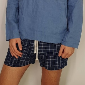 Op de afbeelding: Een blauw linnen shirt met een knoopsluiting en lange mouwen. Het shirt heeft een opgestikte zak aan de voorkant en een trekkoord in de taille. De persoon draagt ​​blauw-wit geblokte shorts.