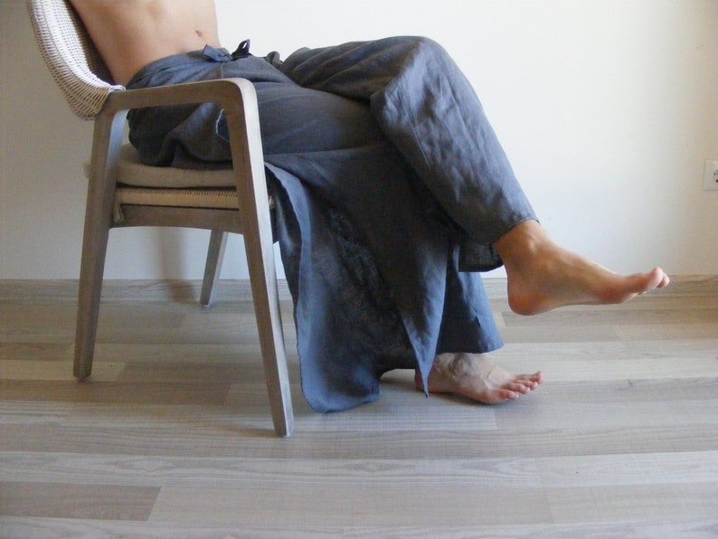May include: A person sitting in a light wood chair with a white cushion. They are wearing loose-fitting, dark gray pants.