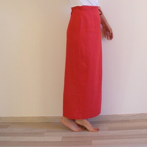 May include: A long, bright red skirt made of linen. The skirt has a simple, straight silhouette and a high waist. It is shown on a model standing on a wooden floor.