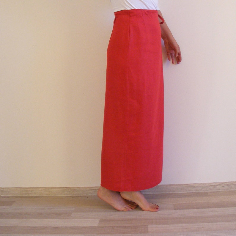 May include: A long, bright red skirt made of linen. The skirt has a simple, straight silhouette and a high waist. It is shown on a model standing on a wooden floor.