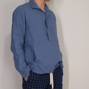 May include: A light blue long-sleeved shirt with a button-up collar and a single button at the neck. The shirt has a large pocket on the front and a drawstring at the waist.