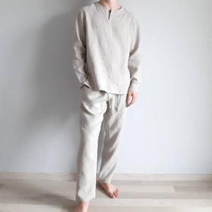 May include: A beige linen pajama set with a long-sleeved shirt and pants. The shirt has a V-neck and a button closure at the neckline. The pants have a drawstring waistband.