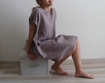 May include: A woman wearing a loose-fitting, light brown linen dress. The dress has a simple, rounded neckline and short sleeves. She is sitting on a small, gray box.