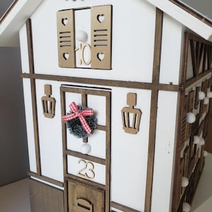 May include: A wooden advent calendar in the shape of a house, painted white with brown trim. The house features a door with the number 23, a wreath, and a mailbox. The windows have shutters and the number 10. Small white lights are attached to the side.