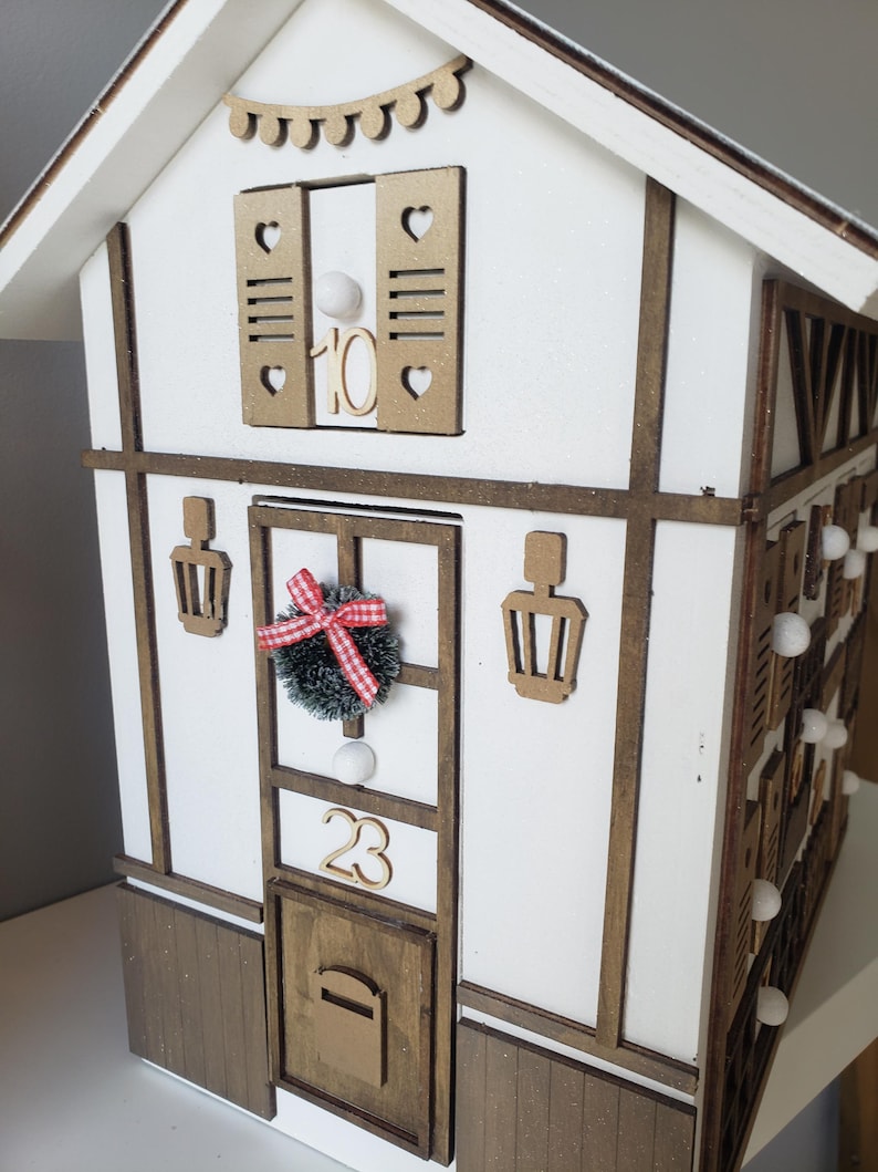 May include: A wooden advent calendar in the shape of a house, painted white with brown trim. The house features a door with the number 23, a wreath, and a mailbox. The windows have shutters and the number 10. Small white lights are attached to the side.