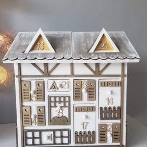 May include: A white wooden advent calendar shaped like a house, with small drawers and doors numbered 1 to 24. The roof is grey with wooden trim, and the house has a rustic, farmhouse style.