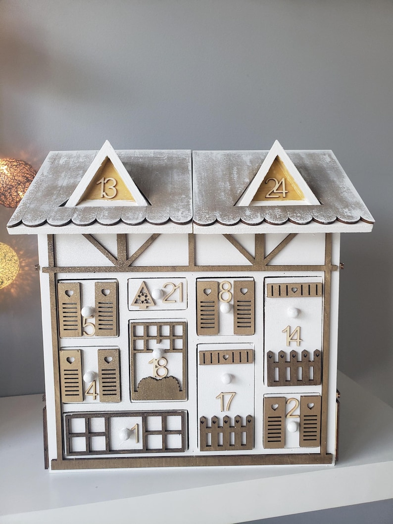 May include: A white wooden advent calendar shaped like a house, with small drawers and doors numbered 1 to 24. The roof is grey with wooden trim, and the house has a rustic, farmhouse style.