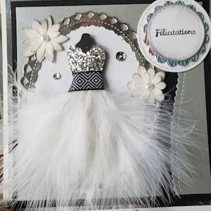 May include: A white card with a black and silver border. The card features a white feather boa with a silver and black sequined dress in the center. The card also has a white circle with the word "Felicitation" in black text.