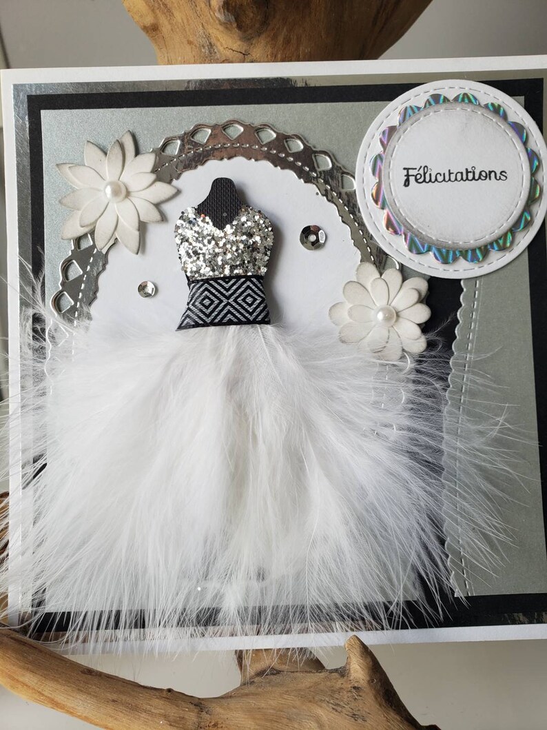 May include: A white card with a black and silver border. The card features a white feather boa with a silver and black sequined dress in the center. The card also has a white circle with the word "Felicitation" in black text.
