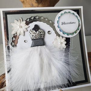 May include: A white card with a silver glitter dress, white feathers, and a white circle with the word "Felicitation" in black text.