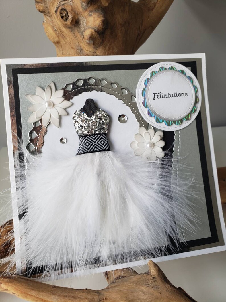 May include: A white card with a silver glitter dress, white feathers, and a white circle with the word "Felicitation" in black text.
