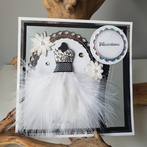 May include: A white card with a black and silver border. The card features a white feather dress with a black and white patterned bodice. The dress is adorned with silver glitter and rhinestones. A white flower with a pearl center is on either side of the dress. A circle with a scalloped edge and the word "Felicitation" in rainbow lettering is in the upper right corner.