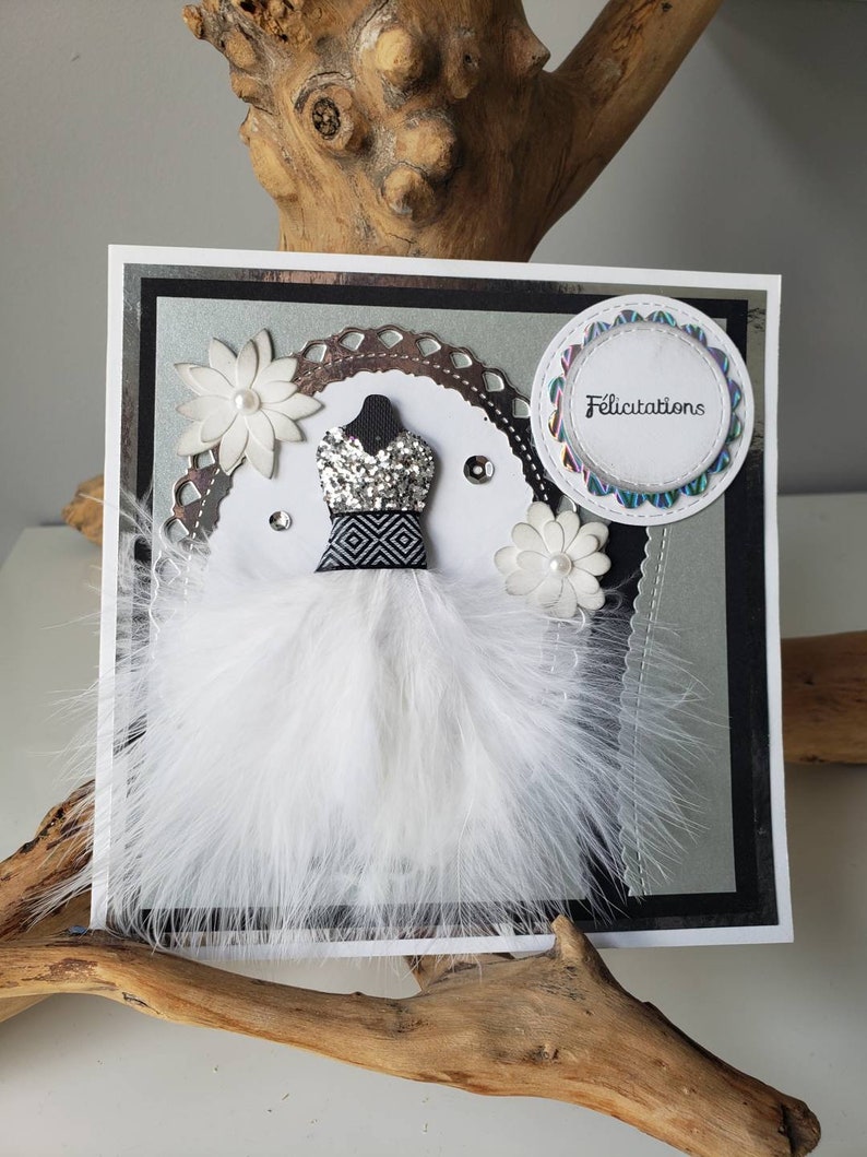 May include: A white card with a black and silver border. The card features a white feather dress with a black and white patterned bodice. The dress is adorned with silver glitter and rhinestones. A white flower with a pearl center is on either side of the dress. A circle with a scalloped edge and the word "Felicitation" in rainbow lettering is in the upper right corner.