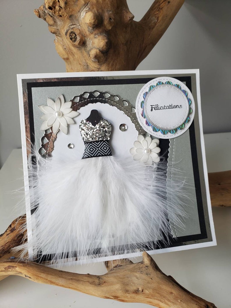 May include: A white card with a silver and black border. The card features a white feather boa, a silver and black dress, and a white flower. The card also has a circle with the word "Felicitation" in black text.