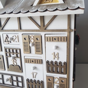 May include: A white wooden advent calendar in the shape of a house, featuring a gray roof and gold detailing. The 24 doors are individually numbered and designed. Small white pom-poms add to the festive decoration.