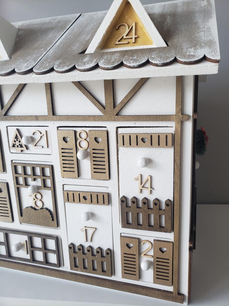 May include: A white wooden advent calendar in the shape of a house, featuring a gray roof and gold detailing. The 24 doors are individually numbered and designed. Small white pom-poms add to the festive decoration.